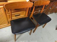 Two teak wood chairs with black faux leather seats, showing wear and scratches on seats and wood frames
