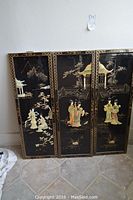 Three rectangular Asian decorative panels with black backgrounds and gold-painted intricate scenes including figures, pagodas, trees, and mountains. Panels display wear and scratches.
