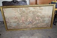 Full view of large framed tapestry showing pastoral scene with several figures and animals by river and trees in muted earth tones.