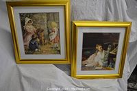 Two gold-framed prints standing against a white cloth background showing children and a dog in outdoor scenes.
