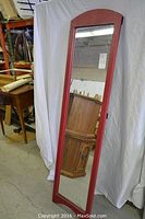 Full view of the tall, narrow mirror with red faux leather frame standing upright against a white backdrop in a warehouse setting.