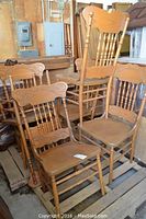 Six oak chairs shown in stacked arrangement with visible carved backs and spindled legs. Chairs show surface scratches and wood wear.