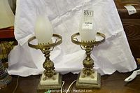 Pair of metal and marble lamps with frosted glass chimney shades on a dark wood floor against a white backdrop.
