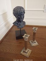 Image showing two silver-tone decorative candlesticks with Corinthian capitals next to a classical-style bust sculpture on black rectangular base, positioned on hardwood floor