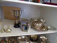 Shelf view of multiple silverplate pieces including trophy, salt and pepper, lidded dish, trays and tea caddy