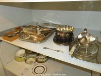 Photo showing assorted silverplate servingware items on shelf including trays, lidded entree dish, small stacked bowls, and cheese board.