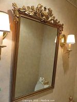 Full view of the rectangular decorative wall mirror with gold leaf decorative urn and leaf crest top, mounted on beige wall with sconces.