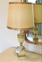 Full view of one bedside lamp on table with cream-colored drum shade and gold trim, urn-shaped body, and square base, reflecting in mirror behind