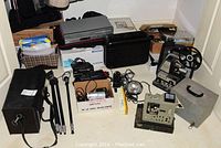 Photo showing multiple vintage electronics items including Bell & Howell projector, Eumig projector, tripods, film reel, and various accessories arranged on a floor area.