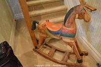 Side angle view of colorful wooden rocking horse showing its painted saddle and carved details near staircase.