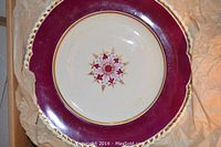 Top view of a porcelain dinner plate showing a purple and pink floral design in the center with a deep maroon border and gold accents around the edge.