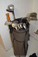 Photo showing golf clubs standing in golf bag, including irons and woods, umbrella handle visible.