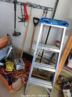 Photo showing the Reynolds aluminum ladder with blue top tray, adjacent tool rack wall with hand tools, battery charger with orange extension cords, and wooden snow shovel.
