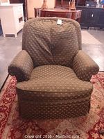 Front view of one green upholstered swivel armchair showing broad rounded arms, cushioned backrest, and fabric skirt base on a patterned rug.