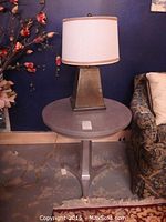 Round grey painted pedestal side table with brass accent metal feet, 24 in. diameter x 24 in. high, next to sofa and floral decoration, with metal base table lamp placed on top.