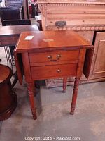 Full view of rectangular drop leaf table with two drawers, two turned legs in front and drop leaf sides extended, reddish-brown wood.
