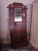 Full front view of antique wooden hall tree with beveled glass mirror, small drawer beneath mirror, open lower compartment and six metal coat hooks.