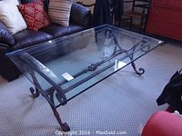 Full view of clear rectangular glass coffee table on metal scroll legs in room with carpet and furniture.