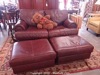 Full view of burgundy red leather loveseat with nailhead trim on rolled arms and two matching leather ottomans in front, placed on decorative rug.