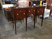 Full view of Kittinger console showing three drawers and tapered legs on a concrete floor in a warehouse.