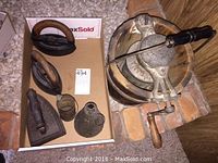 Photo showing three vintage flat irons with wooden handles and two tin pitchers on cardboard surface.