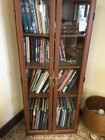 Front view of wooden bookcase with double glass doors showcasing the arranged books inside. The bookcase is medium-brown wood with brass handles and clear glass panes.