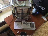 Full front view of the JukeMaster 100 chrome decorative jukebox on a table next to the manuals and adapter.