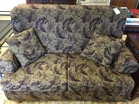 Front view of loveseat with two square matching pillows, showing floral upholstery with muted green, purple, and beige tones.