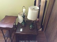 Pair of green ceramic table lamps with white fabric shades (one shade detached) on top of a small wooden cabinet beside wooden decorative shelf.