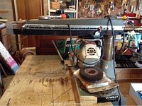 Full side view of Black & Decker radial arm saw on metal stand with wooden top and miscellaneous workshop items around.