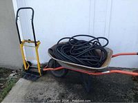 Wheelbarrow with garden hose coiled inside and yellow dolly standing beside it against a white wall.