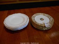 Two stacks of plates on wooden surface: left stack of 5 white Copelands plates with scalloped gold trim, right stack of 8 decorative Hammersley plates with gold floral border and floral center.