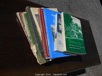 Photo shows six local history books stacked on a dark wooden surface. Books have worn and torn covers. The top book is titled 'Homesteads' with a green and white cover showing a historic house image.
