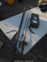 Fishing rods, tackle box, fish scaler displayed on a white table outdoors on gravel ground.
