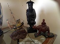 Three Asian wooden carvings on a glass table: fisherman with fishing rod, water buffalo with boy riding, and fishing boat on rock base.