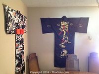 Full view of two silk kimonos hanging on wall, showing black kimono with gold dragon embroidery and black kimono with white crane and floral patterns with red obi.