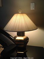 Single brass table lamp with pleated beige shade illuminated, showing the lamp's design details and light effect.