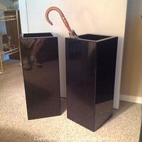 Two black acrylic rectangular umbrella stands shown side by side, with one holding a wooden handled umbrella.