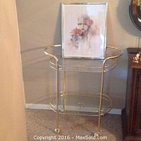 Metal and glass liquor cart with two glass shelves and gold-tone metal frame, on carpeted floor near brown wall.