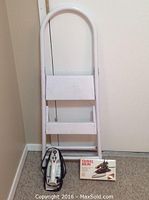 Photo of white metal step stool with two steps and curved handle, alongside electric iron and boxed travel iron on floor.