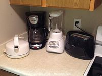 Photo showing four small kitchen appliances on a beige countertop: white electric kettle, black drip coffee maker, white Osterizer blender, and black 2-slice toaster.