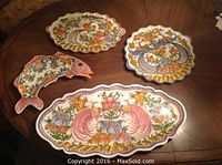 Four handmade Greek pottery serving platters displayed on a wooden table; three oval and one fish-shaped, all with vibrant floral and bird designs.