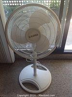 Front view of white Lasko standing fan with three blades and circular grille, placed on carpeted floor near window.