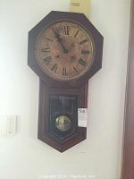 Full view of the regulator wall clock mounted on a wall showing upper octagonal face and lower pendulum housing.