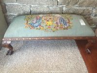 Full view of bench showing carved wood frame and needlepoint upholstered top against a stone wall backdrop.