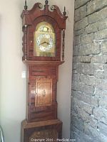 Front view of tall ornate grandfather clock with original wooden case beside stone wall.