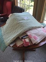 Five handmade quilts folded on a wooden table inside a room with window lighting, showing differing colors and textures