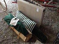 Photo showing beige plastic folding table folded upright with striped cushion and a green bag containing rope hammock or gazebo parts on gravel floor.