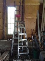 Three ladders, two aluminum step ladders in front and one wood extension ladder behind near a window.