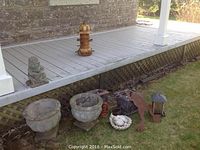 Concrete planters and garden ornaments placed near a porch, showing two large round concrete planters, a concrete child figurine, metal garden ornaments, and a wooden birdhouse on a grass patch near a wooden deck.